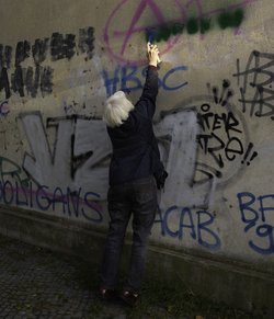 Ältere Frau steht auf Zehenspitzen vor einer Wand und übersprüht eine Hassbotschaft.