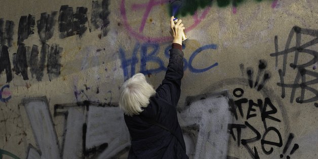 Ältere Frau steht auf Zehenspitzen vor einer Wand und übersprüht eine Hassbotschaft.