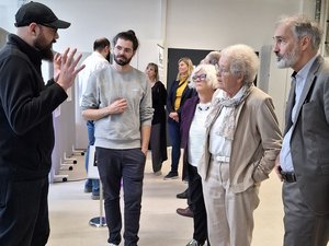 In einem Raum stehen einige Menschen. Sie hören zwei Männern zu, die einen Vortrag halten.