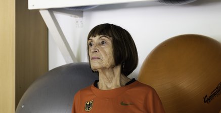 Eine ältere Frau steht selbstbewusst vor einem Regal mit Gymnastikbällen. Sie trägt ein rotes Sportoberteil.