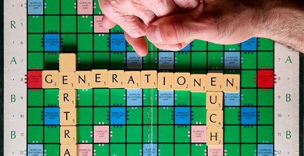 Scrabble-Brett mit den Worten "Generationen vertragt euch". Das V ist blank. 2 altersungleiche Hände berühren sich.
