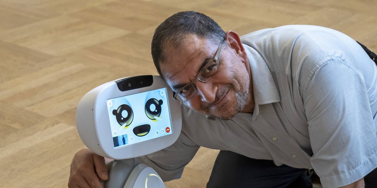 ©BAGSO/Sachs Älterer Mann kniet auf dem Fußboden und legt den Arm um die Schulter eines kleinen Roboters.