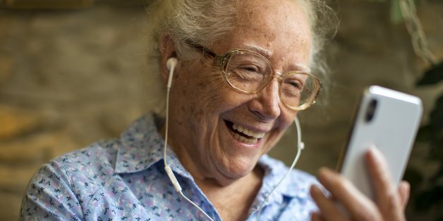 Alte Frau mit Kopfhörern im Ohr schaut lächelnd auf ihr Smartphone.