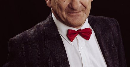 Ein älterer Mann mit zurück gekämmten Haaren vor schwarzem Hintergrund mit Hemd und roter Fliege.