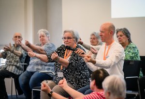 Ältere Menschen sitzen in einem Veranstaltungsraum auf Stühlen. Gerade strecken sie alle ihre Hände nach vorne. In der Mitte hält ein älterer Mann in weißem Sweatshirt einer ein Mikrofon vor den Mund. Sie trägt eine scharz-weiß bedruckte Bluse und hat ein Namensschild um dem Hals, auf dem steht ErlebnisTanz.