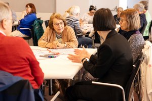Ältere Menschen sitzen um Arbeitstische, die mit Papier bedeckt sind und auf denen Stifte liegen. Im Fokus steht eine ältere Frau  mit beger Jacken, rötlichem Haar und Brille. Sie hört einer Frau, die ihr gegenüber sitzt, aufmerksam zu.