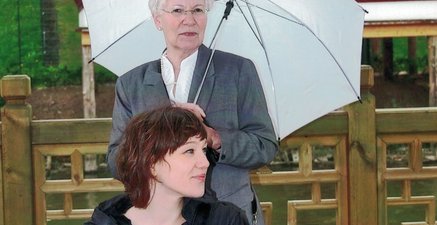 Junge Frau sitzt auf einer roten Bank. Dahinter steht eine ältere Frau mit aufgespanntem Regenschirm.