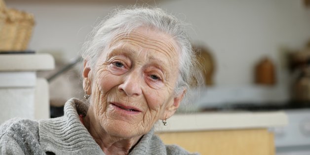 Ältere Frau mit weißem Haar und grauem Strickpullover sitzt in einer gemütlichen Küche und lächelt freundlich in die Kamera.