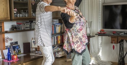 Zwei ältere Frau tanzen lachend und barfüßig vo dem Wohnzimmerschrank
