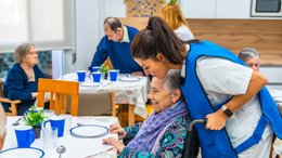 Ein Speisesaal im Seniorenheim. Die Tische sind bereits mit Geschirr gedeckt. Die Bewohnerinnen und Bewohner haben bereits Platz genommen. Ein alte Dame, die im Rollstuhl sitzt, wird gerade von einer Pflegerin an den Tisch gerollt.