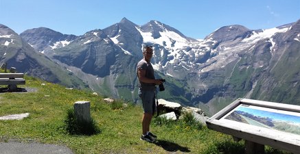 Älterer Mann steht in T-Shirt und kurzer Hose an einem Aussichtspunkt im Gebirge. Er hat eine Fotokamera in der Hand. Im Hintergrund schneebedeckte Gipfel.