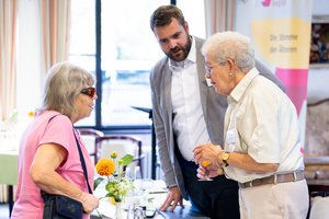 Ein alte Frau mit dunkler Brille, ein junger, großer Mann und eine weitere alte Frau unterhalten sich intensiv.