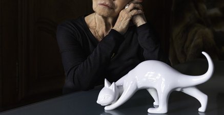 Eine ältere Frau sitzt an einem Tisch, sie blickt nachdenklich zur Seite. Vor ihr steht eine weiße Porzellankatze. 