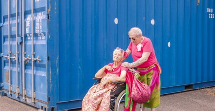 Ein Paar in Urlaubsstyling, vor einem Container. Sie, im Rollstuhl, lächelnd, er hinter ihr, berührt sie liebevoll.
