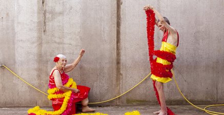 Zwei Senioren vor Betonwand. Sie sitzt, er steht gegenüber. Styling ist aus Lack und Girlanden.        