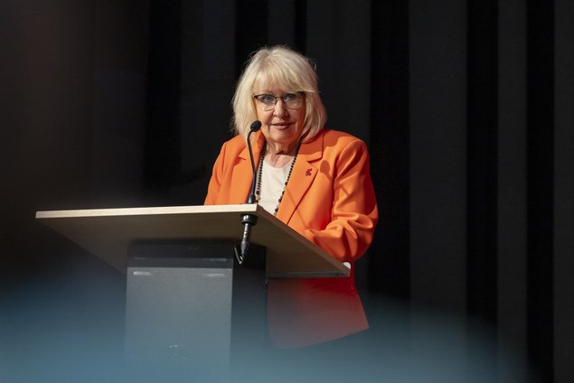 Eine alte Frau in einem orangefarbenen Blazer steht an einem Rednerpult und spricht in ein Mikrofon. Klick öffnet eine vergrößerte Ansicht.