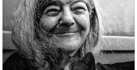 Ältere Frau mit geflochtenen Zöpfen lächelt. Auf ihrem Schoss eine kleine Katze.