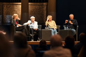 Auf der Bühne: 4 Personen, zwei ältere Frauen, eine Frau mittleren Alters, und ein älterer Mann sitzen in schwarzen Sesseln. Der Mann spricht gerade in ein Mikrofon. 