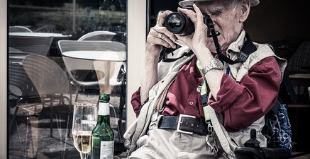 Auf dem Foto ist ein alter Mann zu sehen, der an einem Tisch sitzt und konzentriert durch seine Kamera blickt.