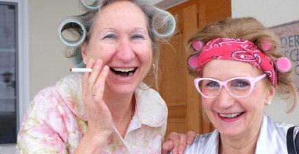 Zwei ältere Damen mit Lockenwicklern lachen. Die eine mit Zigarette in der Hand, die andere mit Brille auf der Nase.