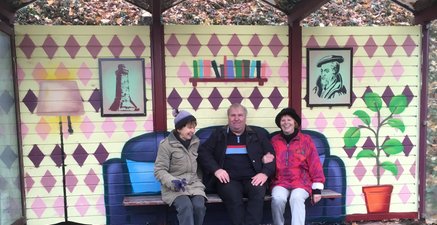 3 Senioren sitzen in einem Buswartehäuschen, die Hinterwand als Wohnzimmer gestaltet