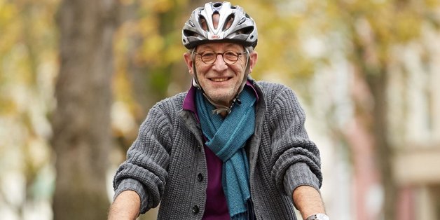 Ein älterer Mann mit Strickjacke, Schal und Helm fährt Fahrrad in der Stadt.