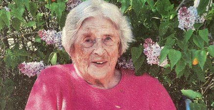 Eine ältere Frau in der Frühlingszeit sitzt mit rosa Pullover neben Fliederbaun mit Blumen und lächelt