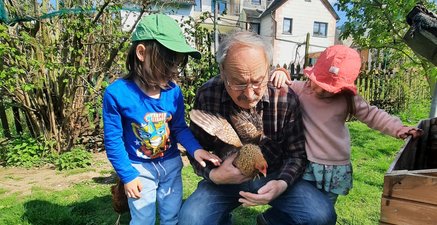 Ein kleines Mädchen und ein Junge stehen bei einem älteren Mann mit Huhn in der Hand.