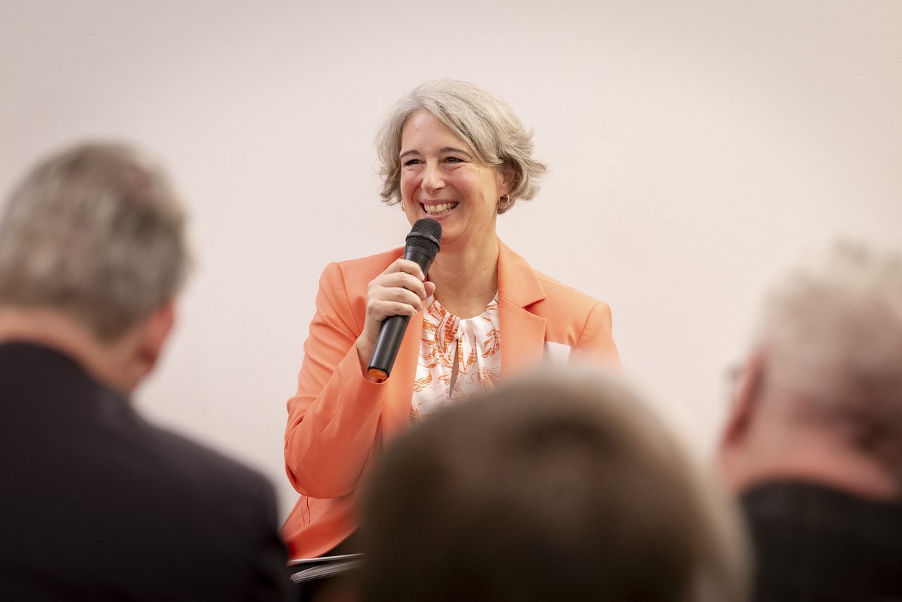 Eine Frau mittleren Alters steht vor Publikum und spricht lächelnd in ein Mikrofon. Sie trägt eine Kurzhaarfrisur und einen orangefarbenen Blazer, darunter eine bunte Bluse. Klick öffnet eine vergrößerte Ansicht.