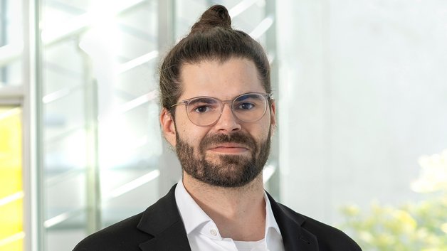 Portrait Alexander Engel: Junger Mann, mit Brille und Bart. Er hat seine schwarzen langen Haare zu einem Knoten hochgebunden. Er trägt ein weißes Hemd und ein schwarzes Sakko.