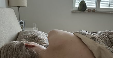 Der Rücken einer Frau im Bett. Der Wecker steht auf kurz vor 12 Mittag. Durch das Rollo scheint die Sonne ins Zimmer.