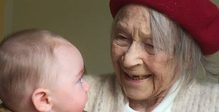 Alte Frau rechts im Bild lacht strahlend einen Säugling an. Die Gesichter sind zueinander gerichtet.