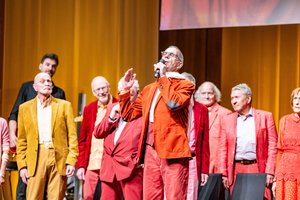 Auf der Bühne: Im Vordergrund ist ein alter Mann zu sehen. Im Anzug in Rottönen und einem weißen Schal um den Hals. Er singt mit geschlossenen Augen in ein Mikrofon. Hinter ihm ist ein Chor zu sehen. Auch in Rottönen gekleidet. Sie schauen auf ihn.