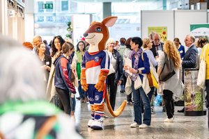 Ein Blick ins Messegeschehen. Viele Menschen stehen in unterschiedlichen Gruppen zusammen und unterhalten sich. Im Vordergrund eine Person in einem Kanguruhkostüm. Sie trägt eine Sporttrikot und eine kurze Sporthose in blau-orange. Die Figur überragt alle.