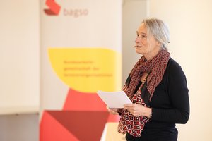 The moderator, Katja Nellissen, a middle-aged woman, is standing on a stage. She is wearing a black sweater, a colourful scarf around her neck and a headset. She is holding notes in her hands. In the background, there is a BAGSO banner.
