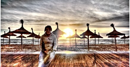 Ältere Frau sitzt am Strand. Hinter ihr stehen Schirme und die Sonne geht unter.
