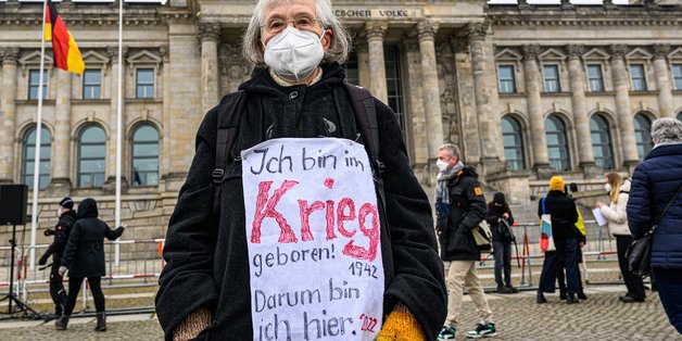 Eine alte Frau steht vor dem Bundestag mit einem Schild vor ihrem Bauch auf dem steht: Ich bin im Krieg geboren! 1942 - Darum bin ich hier. 2022