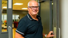 Ein älterer Mann macht gerade eine Tür auf. Im Hintergrund sieht man eine Werkshalle.