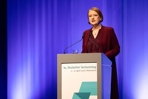 Die Seniorenministerin Lisa Paus, eine Frau mittleren Alters mit rötlichem, schulterlangen Haaren. Sie trägt einen dunkelroten Blazer und eine Bluse. Sie steht am Rednerpult und spricht ins Publikum.