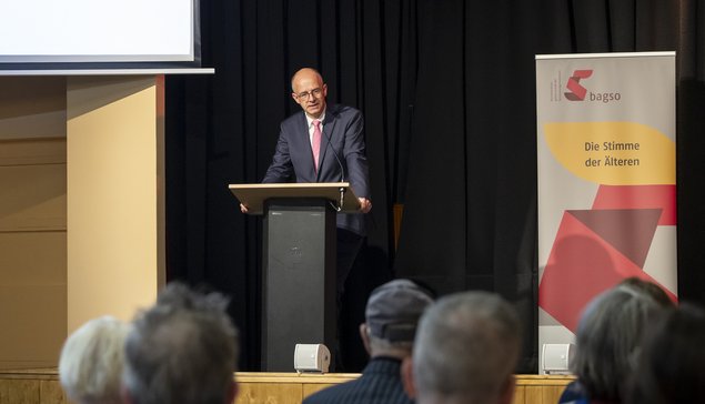 Blick von hinten auf die Bühne. Ein Mann hält an einem Rednerpult eine Ansprache. Oben links auf einer großen Leinwand steht "Wer zuletzt lacht... Willkommen zur Preisverleihung. Klick öffnet eine vergrößerte Ansicht.