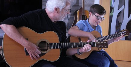 Älterer Mann mit Gitarre zeigt dem neben ihm sitzenden Enkel den passenden Griff