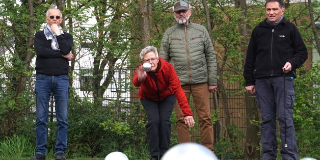 Vier ältere Menschen spielen Bool. Eine Frau in einer roten Jacke wirft gerade die Bool-Kugel. Im Forderung sind die Kugeln auf dem Platz zu sehen.