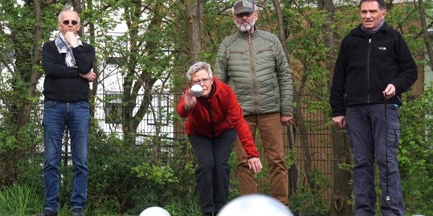 Vier ältere Menschen spielen Bool. Eine Frau in einer roten Jacke wirft gerade die Bool-Kugel. Im Forderung sind die Kugeln auf dem Platz zu sehen.