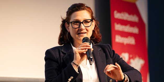 ©BAGSO/Lys Portrait Ferda Ataman: Sie hat dunkles, halblanges Haar, die sie zurückgebunden trägt. Sie hat eine Brille an und ist mit einem schwarzen Blazer und weißer Bluse gekleidet. Sie sitzt auf einem Podium, in einem schwarzen Sessel und spricht in eine Mikrofon.