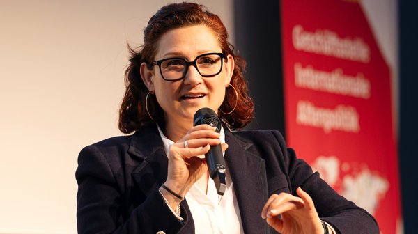 ©BAGSO/Lys Portrait Ferda Ataman: Sie hat dunkles, halblanges Haar, die sie zurückgebunden trägt. Sie hat eine Brille an und ist mit einem schwarzen Blazer und weißer Bluse gekleidet. Sie sitzt auf einem Podium, in einem schwarzen Sessel und spricht in eine Mikrofon.