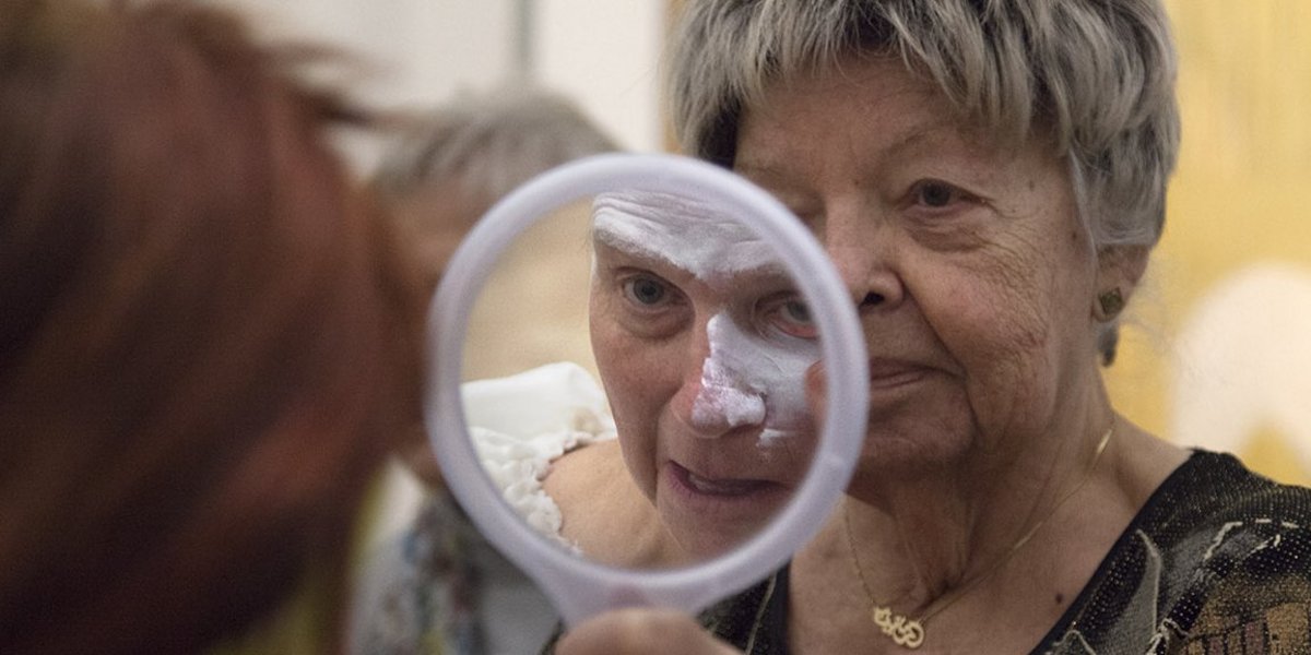 Ältere Frau mit einem Handspiegel, die sie für eine andere ältere Frau hält. Im Spiegelbild ist zu sehen, dass die zweite Frau weiße Schminke auf ihr Gesicht aufträgt.
