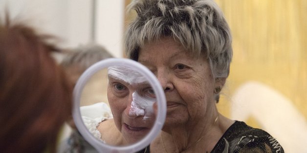 Ältere Frau mit einem Handspiegel, die sie für eine andere ältere Frau hält. Im Spiegelbild ist zu sehen, dass die zweite Frau weiße Schminke auf ihr Gesicht aufträgt.