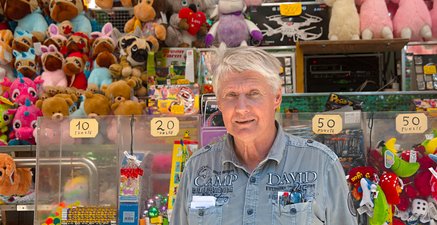 Mann, 68 Jahre, kurze graue Haare, Jeanshemd, steht vor Auslage mit bunten Losgewinnen, Spielzeug, Kuscheltiere