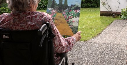 Eine Frau sitzt im Rollstuhl auf der Terrasse und betrachtet ein selbstgemaltes Gartenbild