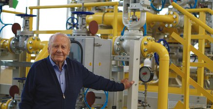 Ein alter Mann steht in einer Werkshalle voller Rohre und Maschinen und schaut stolz in die Kamera.